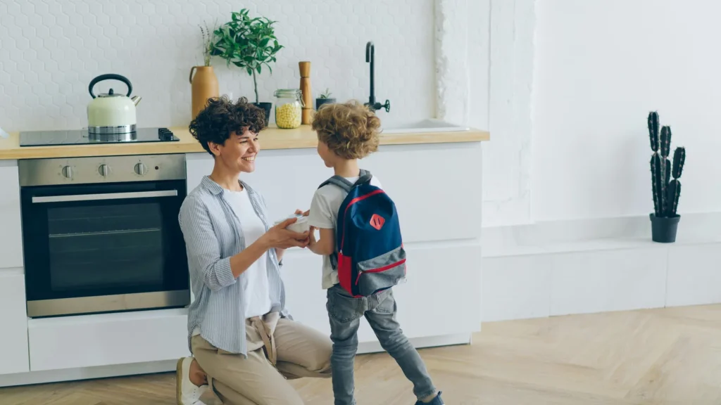 A mum kneeling in the kitchen to help her son with his school backpack